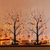 Decorative Halloween setup with bare trees, lit pumpkins, and candles on a table.